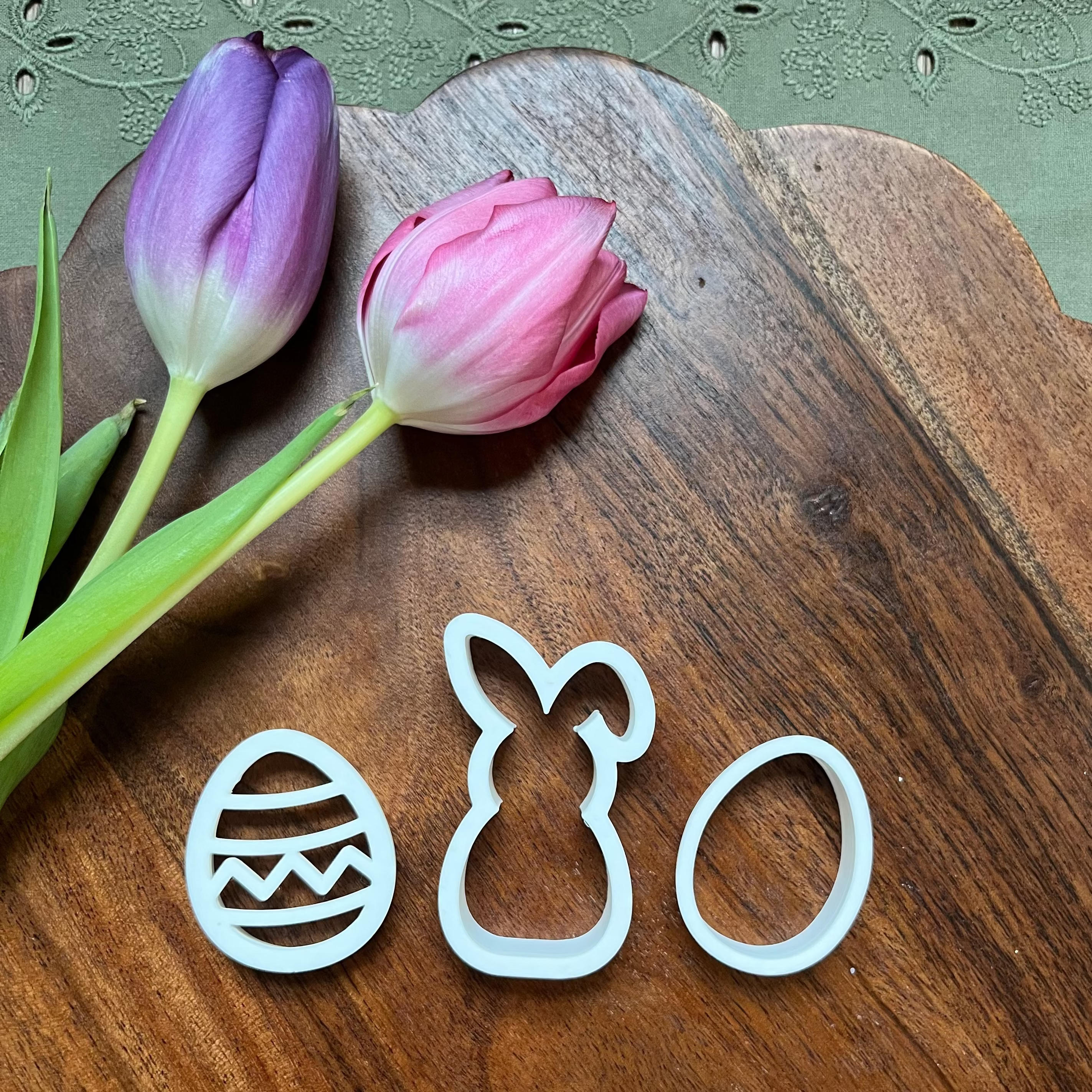 Three cookie cutters shaped like an egg, rabbit, and circle on a wooden surface with tulips.