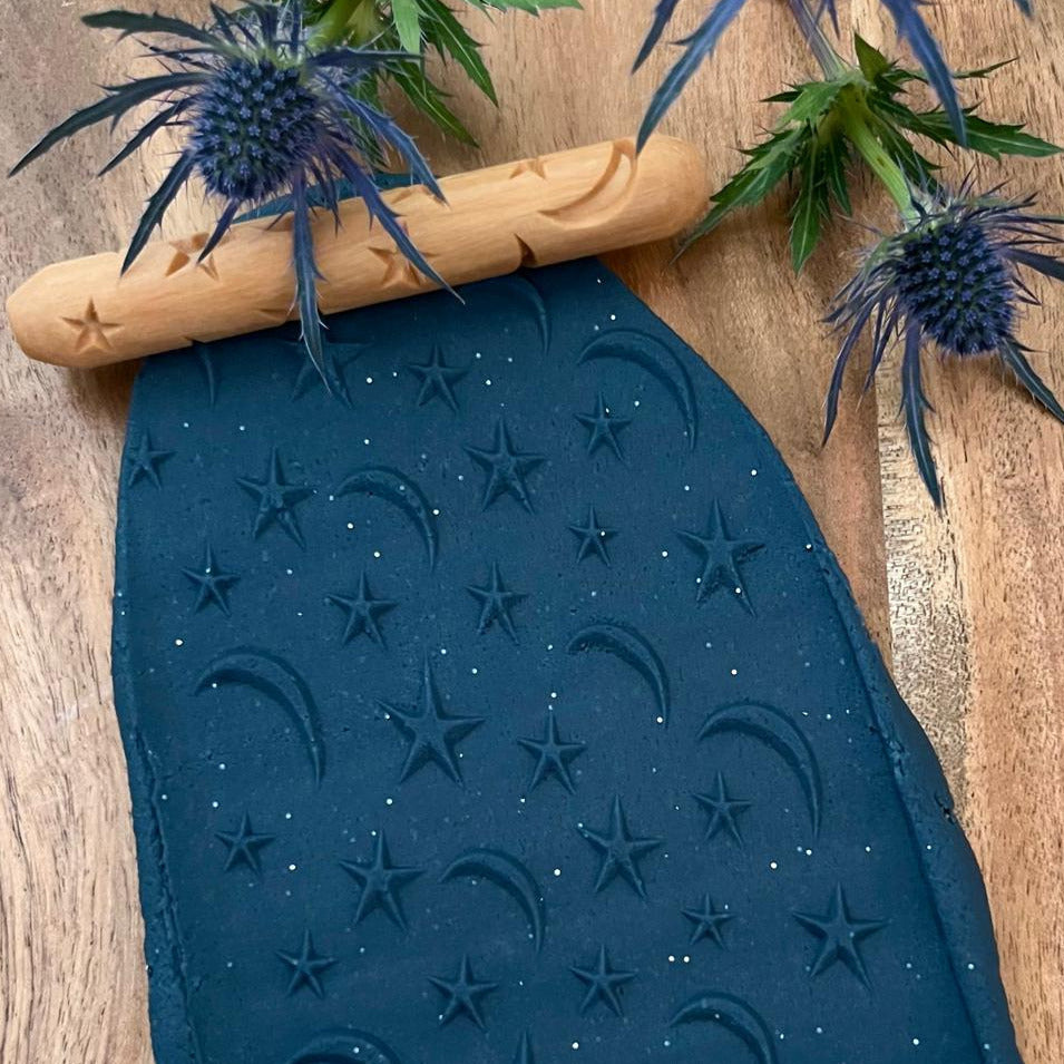 medium sized wooden roller with star and moon engraving rolled along dark blue handmade play dough creating a star and moon pattern.